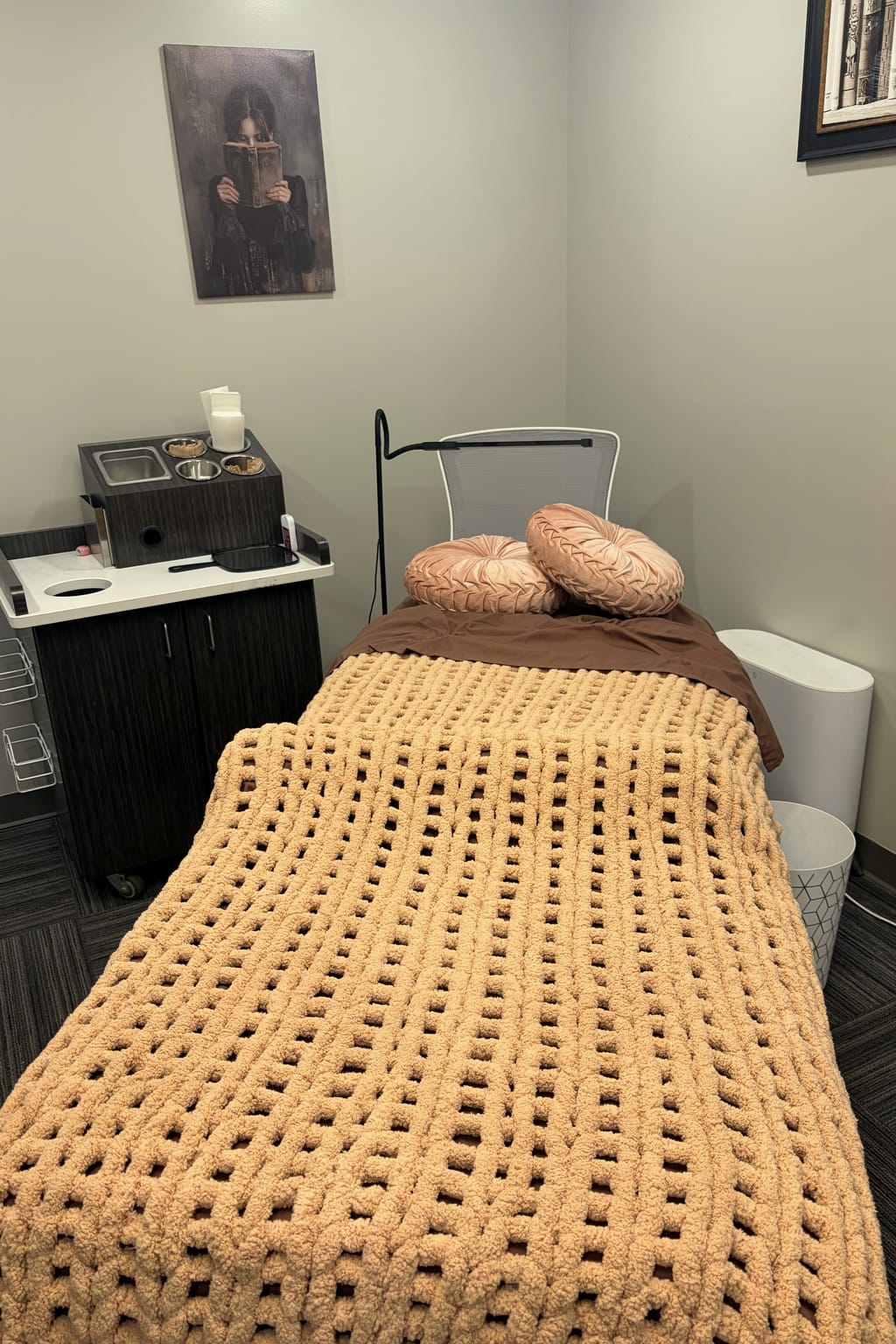 Rachel’s treatment room: a treatment bed with a tan knit blanket and two rose velvet pillows, below a painting of a woman reading a book.